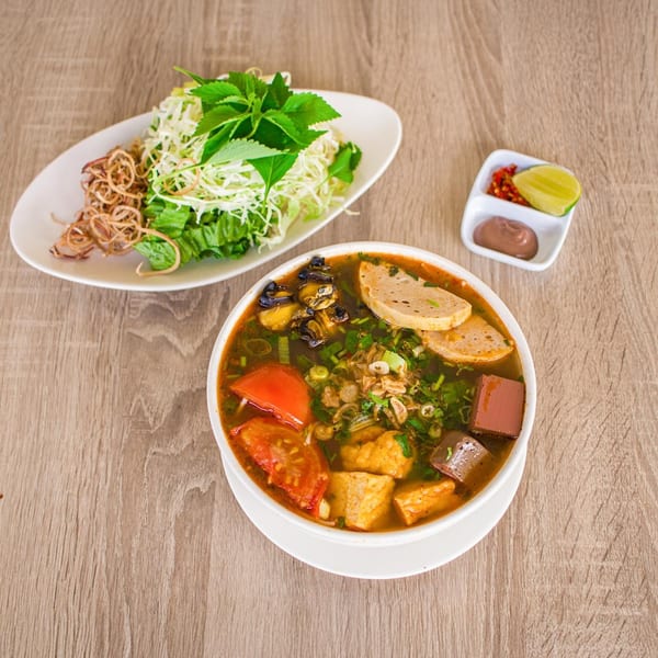 Vietnamese snail noodle soup with fresh herbs, fried shallots, and condiments on a wooden table.