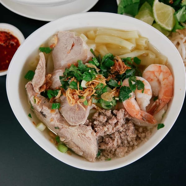A bowl of Vietnamese hu tieu with pork, shrimp, ground meat, and herbs, served with chili sauce and lime.