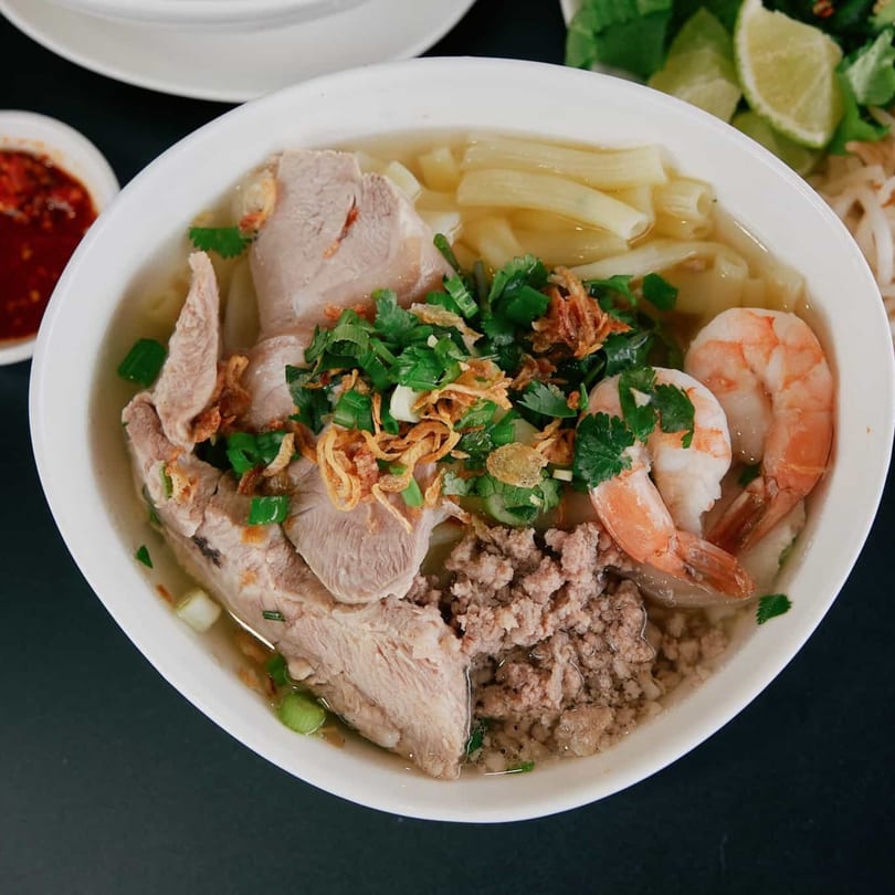 A bowl of Vietnamese hu tieu with pork, shrimp, ground meat, and herbs, served with chili sauce and lime.