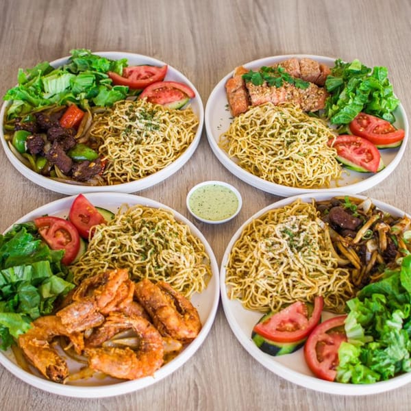 Four Vietnamese noodle dishes with various proteins, fresh greens, tomatoes, and a small bowl of green sauce.