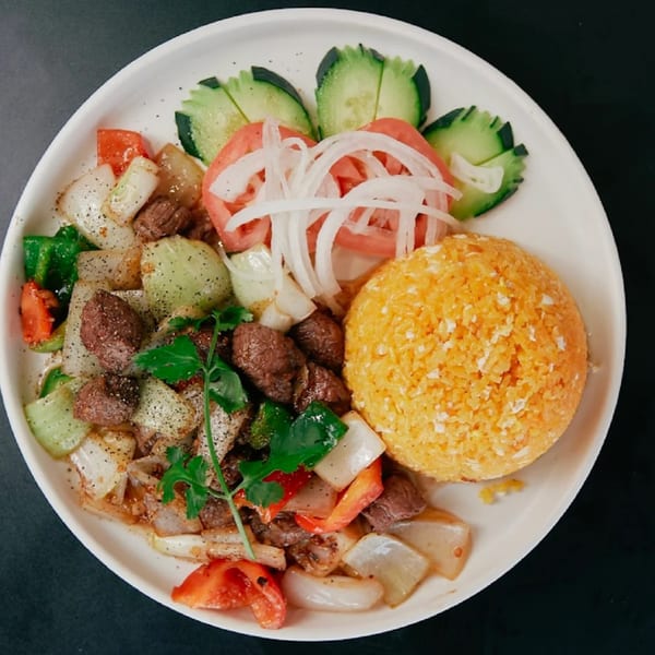 Vietnamese shaken beef with bell peppers, onions, tomatoes, cucumbers, and yellow rice on a white plate.